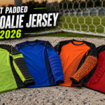 A curated flat-lay display of four distinct, unbranded youth goalie jerseys with various padding technologies, presented on a weathered soccer pitch background for a 2026 buying guide.
