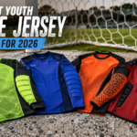 A curated flat-lay display of four distinct, unbranded youth goalie jerseys featuring various padding technologies, presented on a weathered soccer pitch background for a 2026 buying guide.